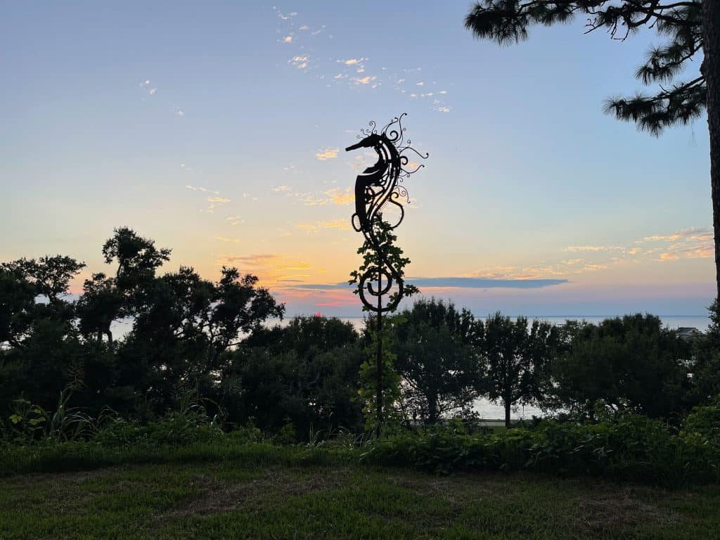 A seahorse sculpture silhouetted against a sunset over Mobile Bay, framed by live oaks